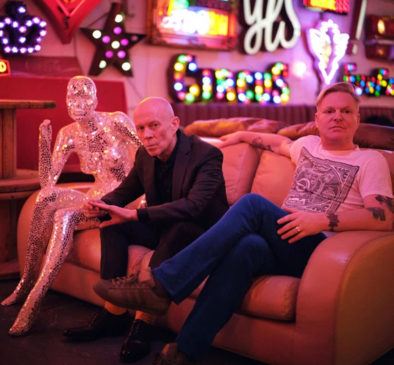 Paul Lavoie and another man sit beside a mirrored mannequin in a lounge filled with colorful neon signs.
