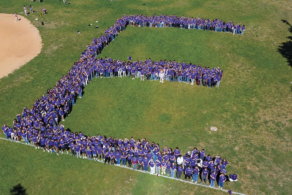 Aerial view of Emerson College students in purple shirts arranged in the shape of a large letter E on a field.