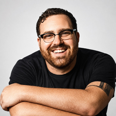 Portrait of James DiSabatino smiling with his arms folded while wearing glasses and a black shirt.