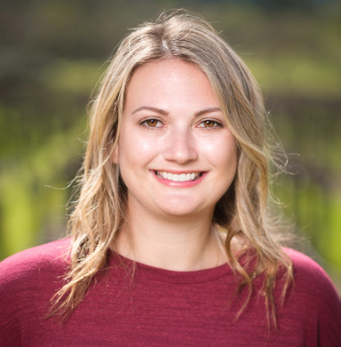 Portrait of Jess Lander smiling outdoors with greenery in the background.