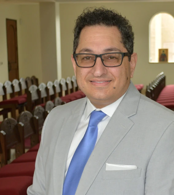 John Khouri smiles in a light gray suit and blue tie inside a church sanctuary.