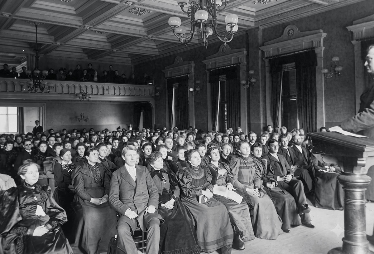 A historical black-and-white photograph of a seated audience listening to a speaker at a podium.