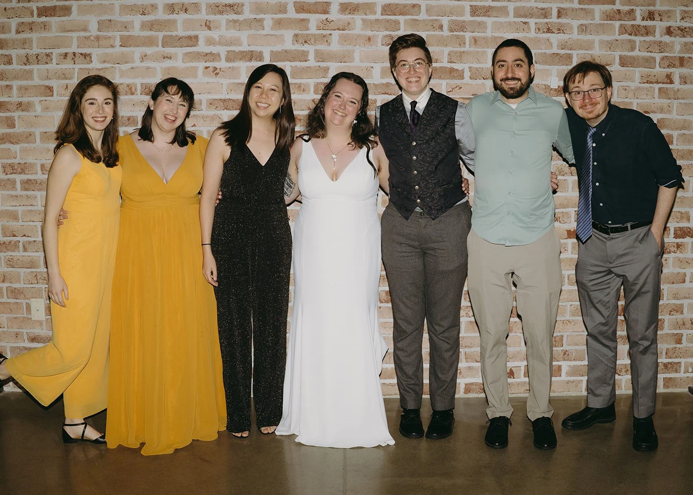 Seven wedding guests pose arm in arm in formal attire against a brick wall.