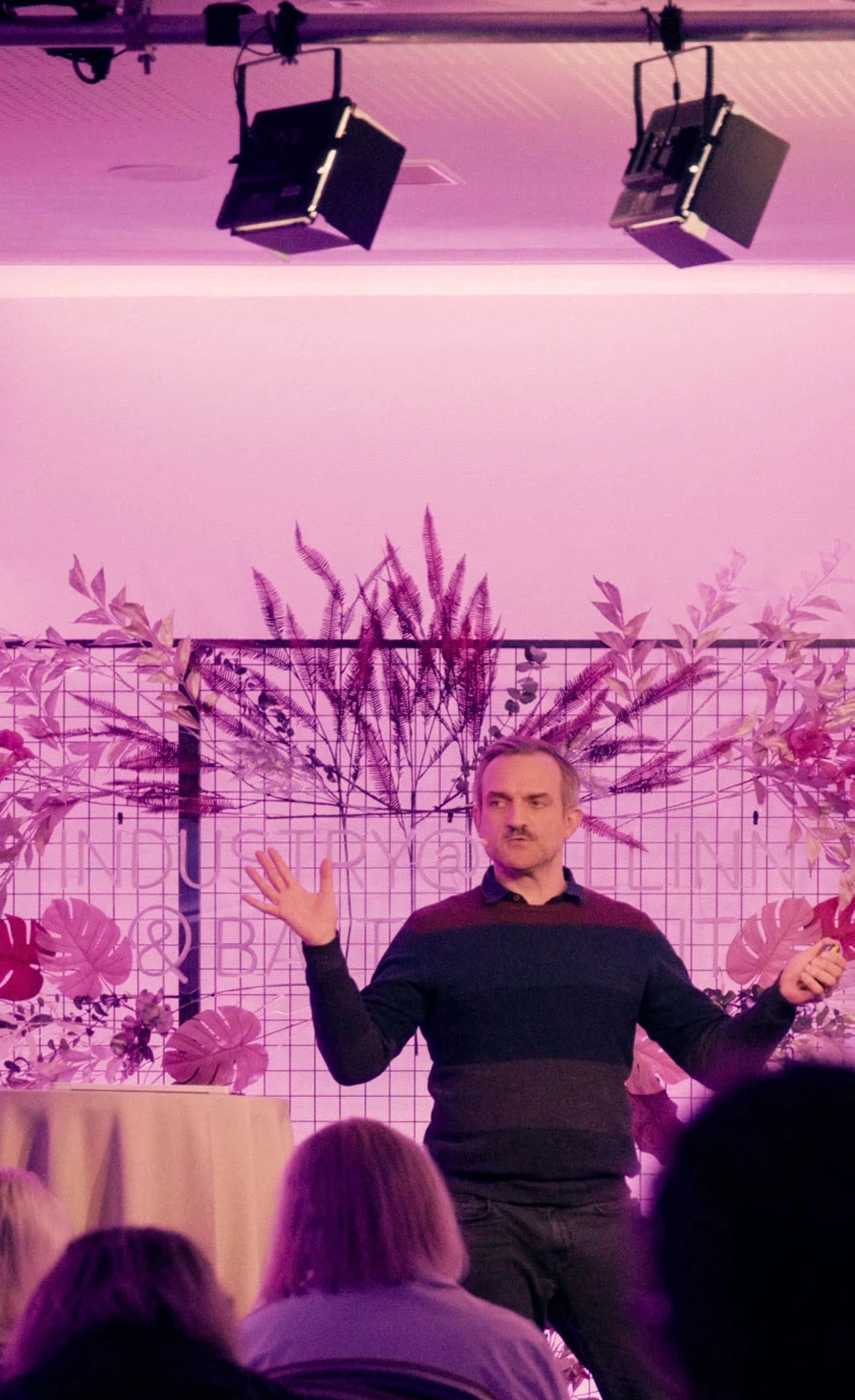 Matt Szymanowski gestures while speaking to an audience on a plant-decorated stage illuminated with purple light.