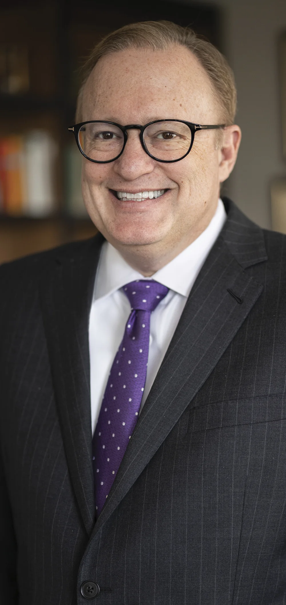 Formal portrait of Emerson College President Jay Bernhardt wearing glasses, a dark suit, and a purple tie.