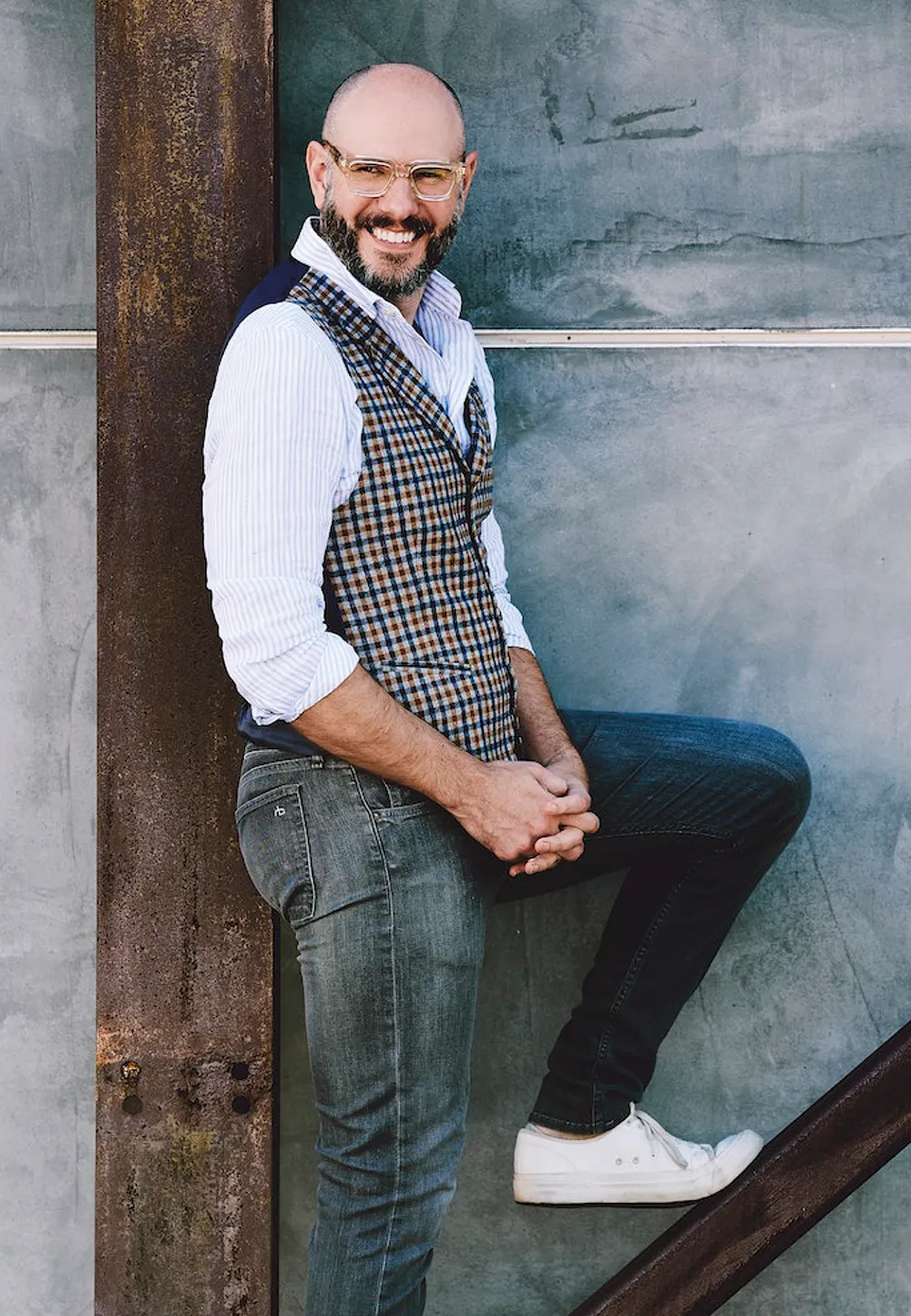 Full-length portrait of Steven Rowley with glasses, beard, striped shirt, and patterned vest against a wall.