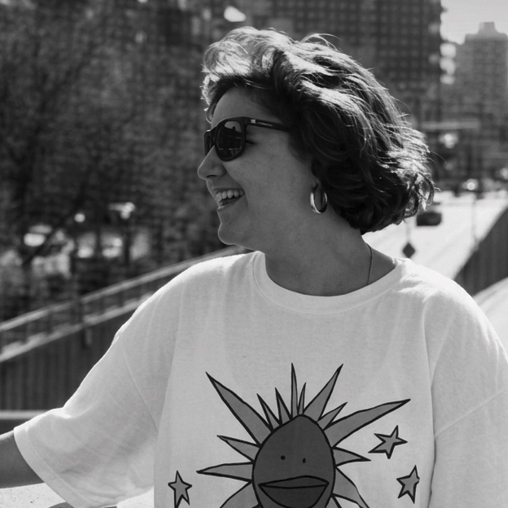 Black-and-white photo of a smiling Holly Bario in sunglasses and a sun-graphic t-shirt outdoors.