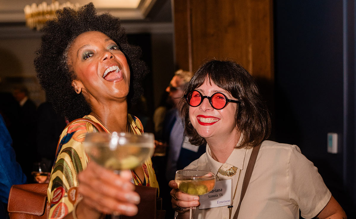 Two women smiling and holding cocktail glasses at an indoor event, one with an afro and patterned outfit and the other wearing red round glasses and a name tag.