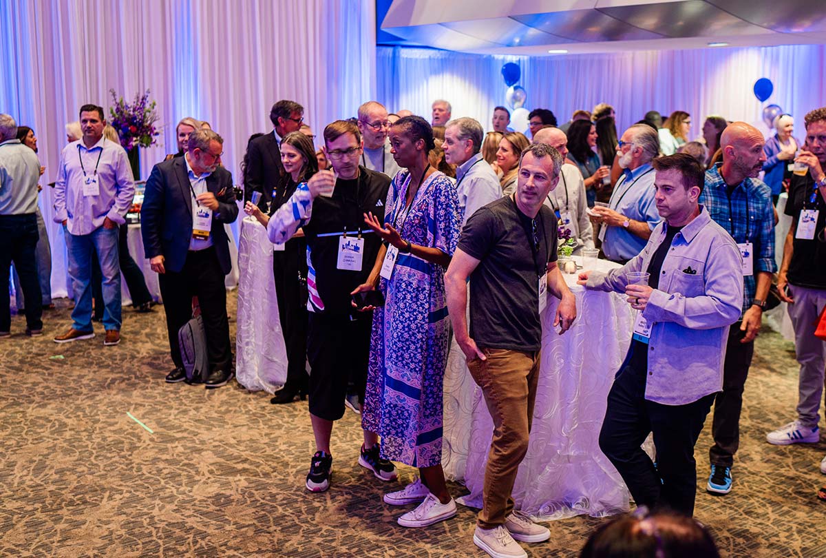 A crowded indoor reception where guests chat and mingle around tall cocktail tables under soft purple and blue lighting.