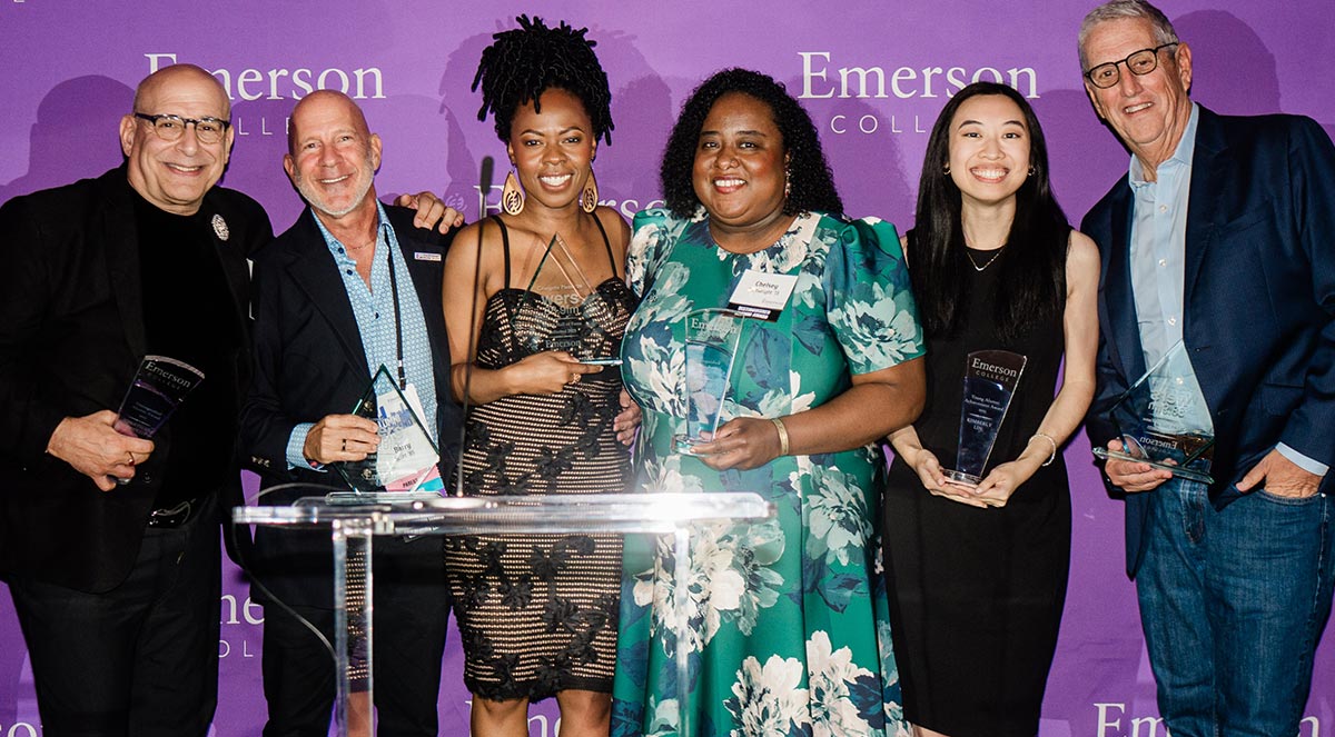 Six award recipients smiling on stage while holding glass trophies in front of a purple Emerson College backdrop.
