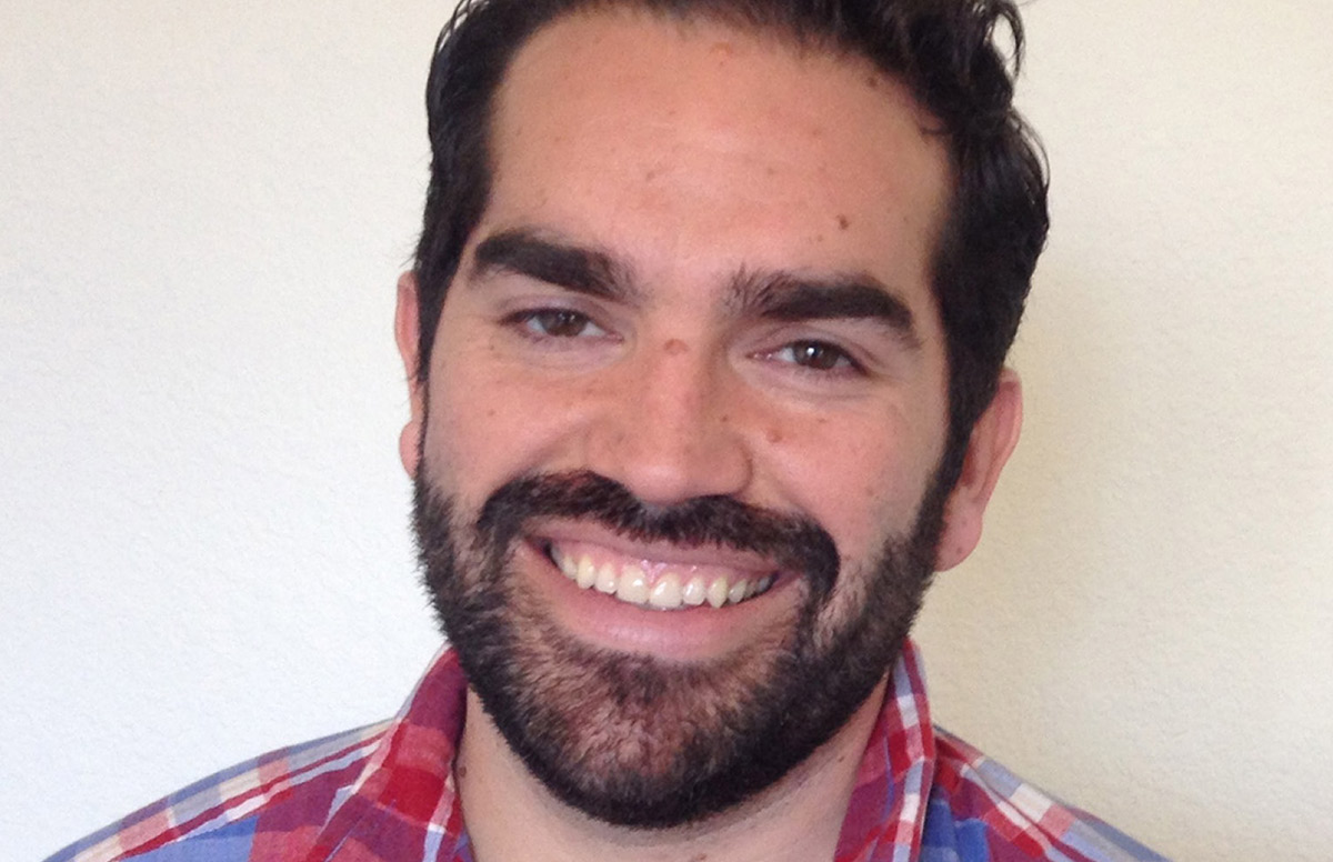 Close-up headshot of a smiling man with dark hair, a full beard, and dark eyebrows, wearing a red and blue plaid shirt.