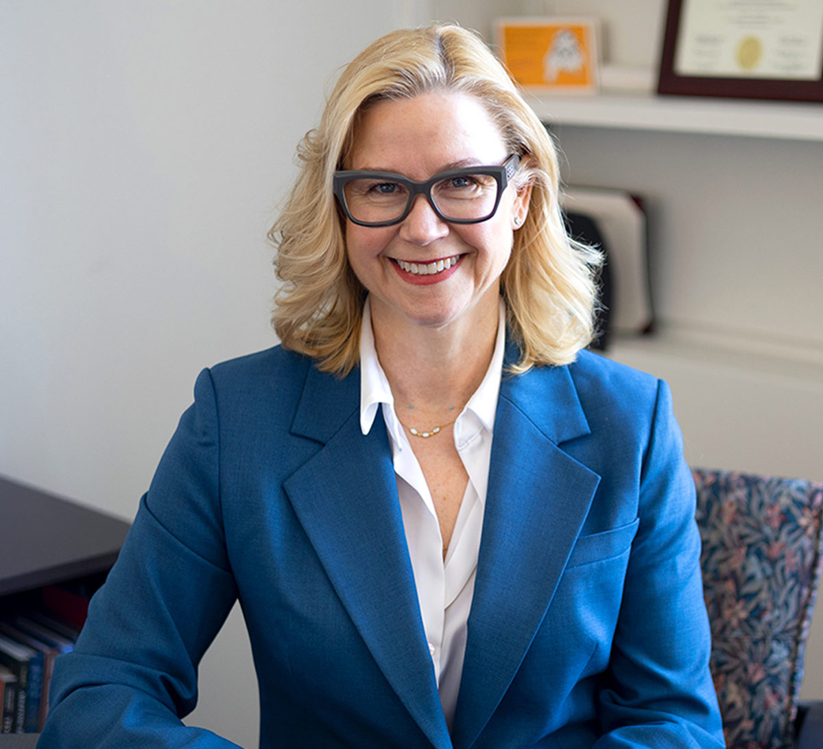 Portrait of a smiling blonde woman wearing dark-framed glasses, a blue blazer over a white collared shirt, seated in an office setting.