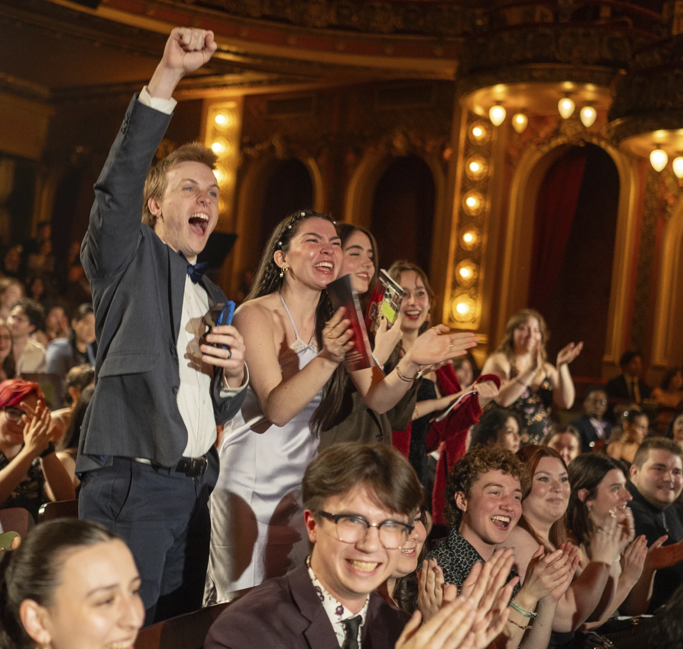 Emerson College students stand, cheer, and applaud during the EVVY Awards ceremony inside a crowded theater.