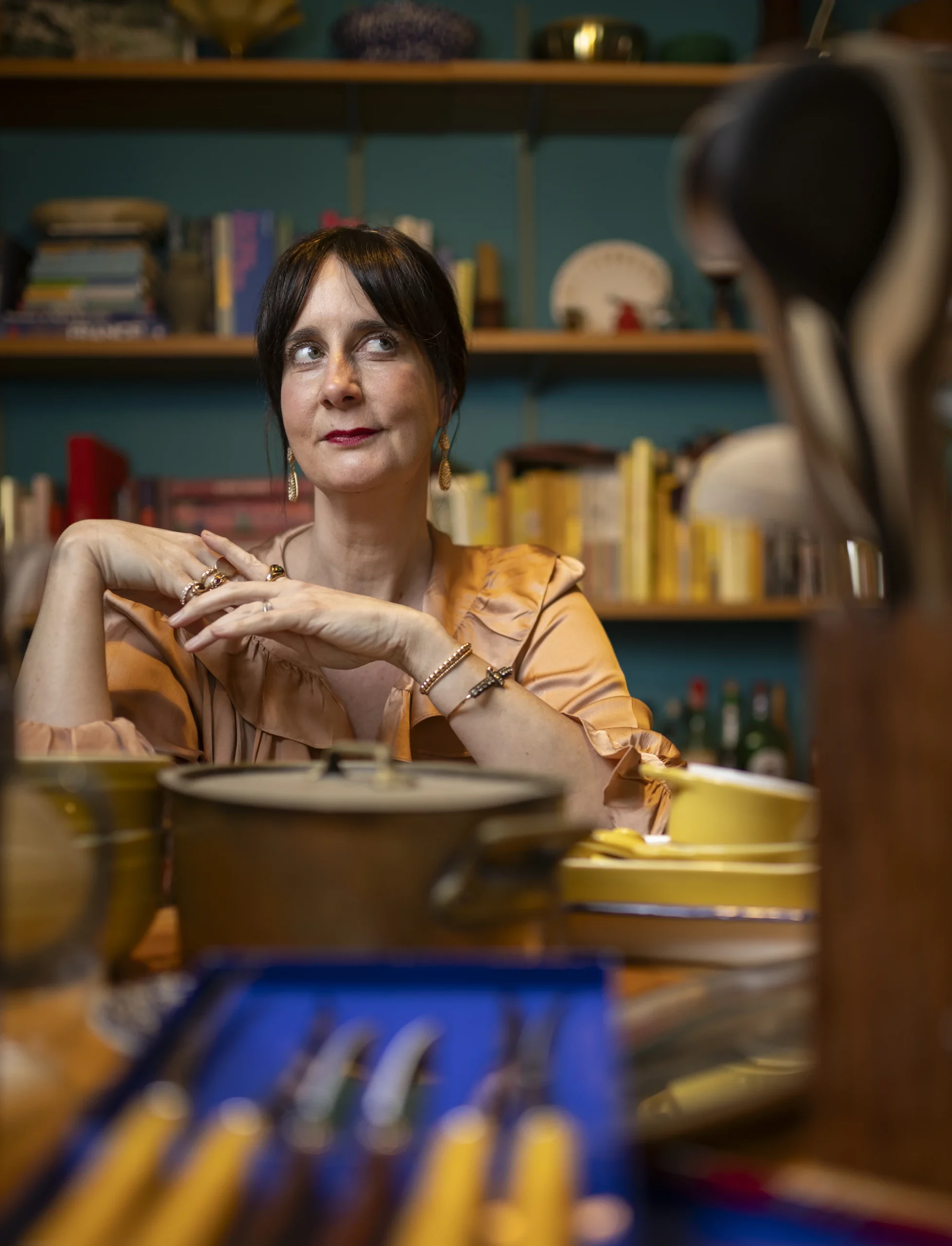 Colu Henry sits at a table surrounded by cookware, with books and kitchen objects displayed on shelves behind her.