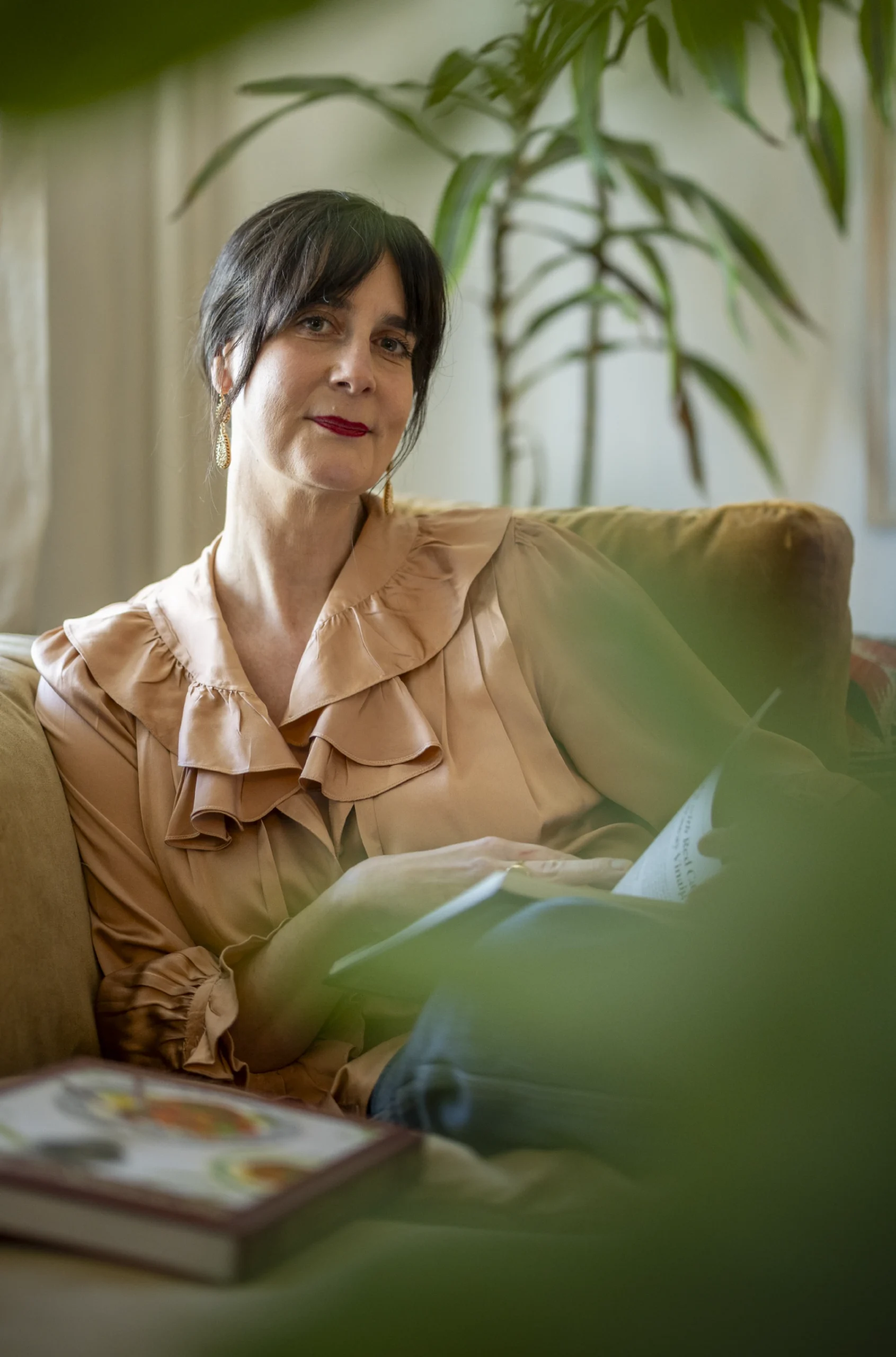 Colu Henry sits on a sofa holding an open book, framed by houseplants in a softly lit room.