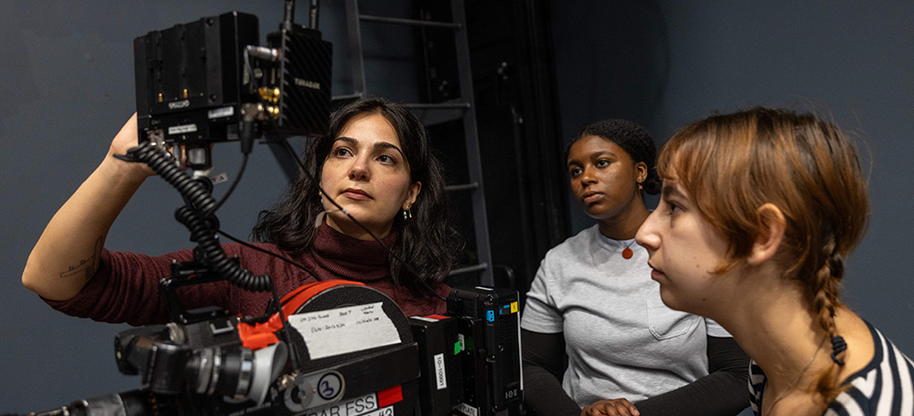 Three film crew members adjusting equipment and focusing intently on a camera monitor.