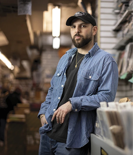 A bearded man in a baseball cap and striped overshirt leans against a counter inside a retail shop.
