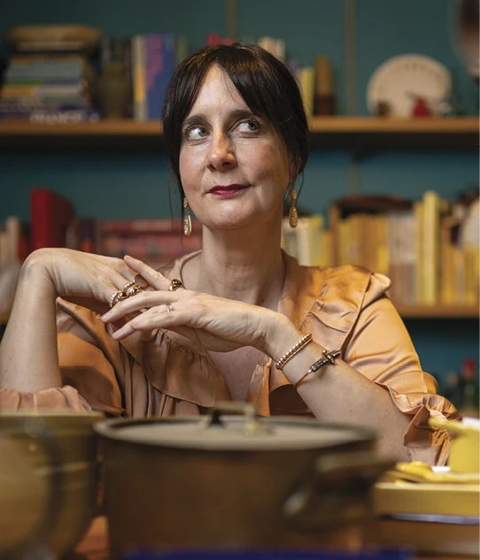 Colu Henry sits at a table with cookware in the foreground and bookshelves behind her.